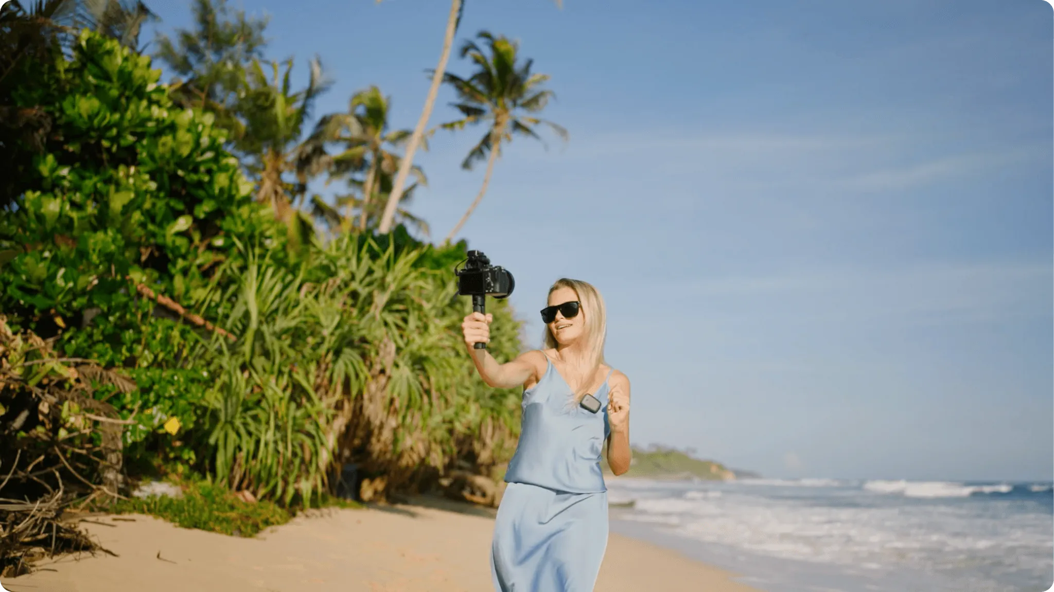 A woman in a blue dress vlogging with a camera and gimbal on a tropical beach.