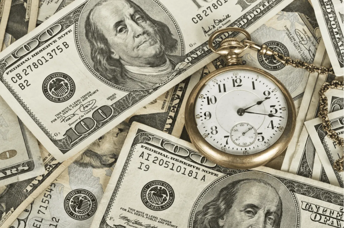 A gold pocket watch rests on US one hundred dollar bills, illustrating 'time is money'.