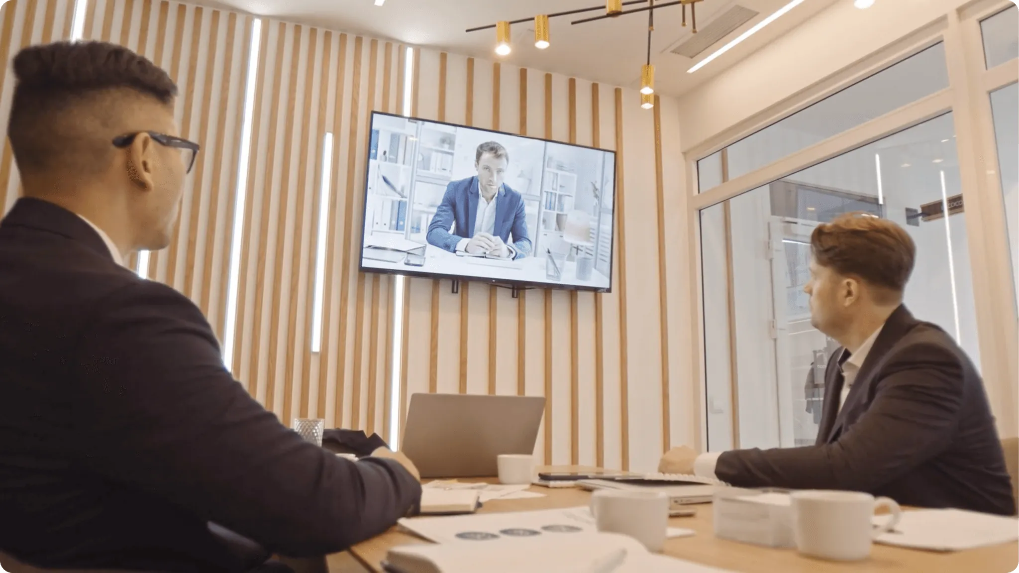 Two men in a modern office meeting room participating in a video call on a large screen.
